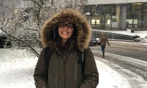 Woman with glasses wearing a parka stands in the snow outside a building at EMBL
