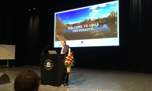 Oliver Billker, Director of MIMS, is standing at the podium in front of a microphone. Behind him, a projected slide says “Welcome to Umeå University”.
